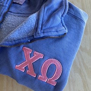 Może przedstawiać: Niebieska bluza z kapturem zapinana na zamek z wyszytymi różowymi greckimi literami X i Ω. Bluza z kapturem ma kaptur ze sznurkiem i zapięcie na zamek. Etykieta marki jest widoczna wewnątrz kaptura.