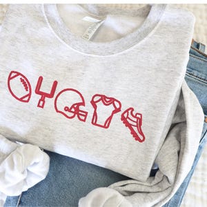 Könnte beinhalten: Ein grauer Pullover mit einem roten gestickten Design aus einem Fußball, einem Torpfosten, einem Helm, einem Trikot und Stollen.