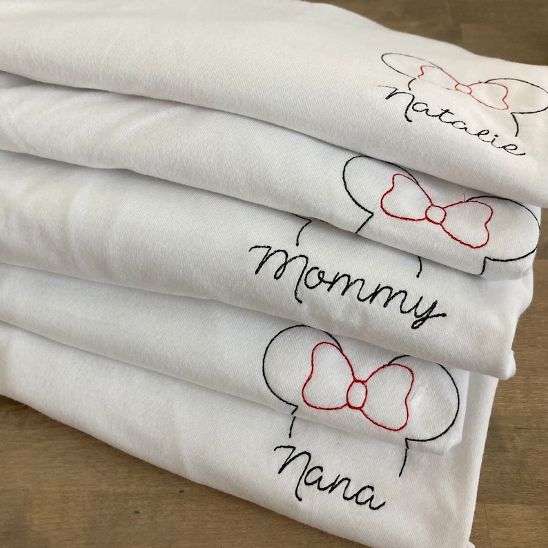 May include: Stack of five white t-shirts with black and red embroidery. The shirts feature a cartoon mouse head with a bow and the names "Natalie", "Mommy", and "Nana" written in cursive.