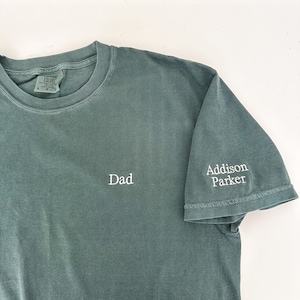 K&ouml;nnte beinhalten: Ein gr&uuml;nes T-Shirt mit dem Wort "Dad" auf der Vorderseite und "Addison Parker" auf dem &Auml;rmel gestickt.