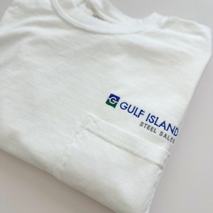 Op de afbeelding: Wit T-shirt met een zak aan de linkerkant. De zak heeft een geborduurd logo met de tekst "GULF ISLAND STEEL SALES" met een groen, blauw en wit logo.