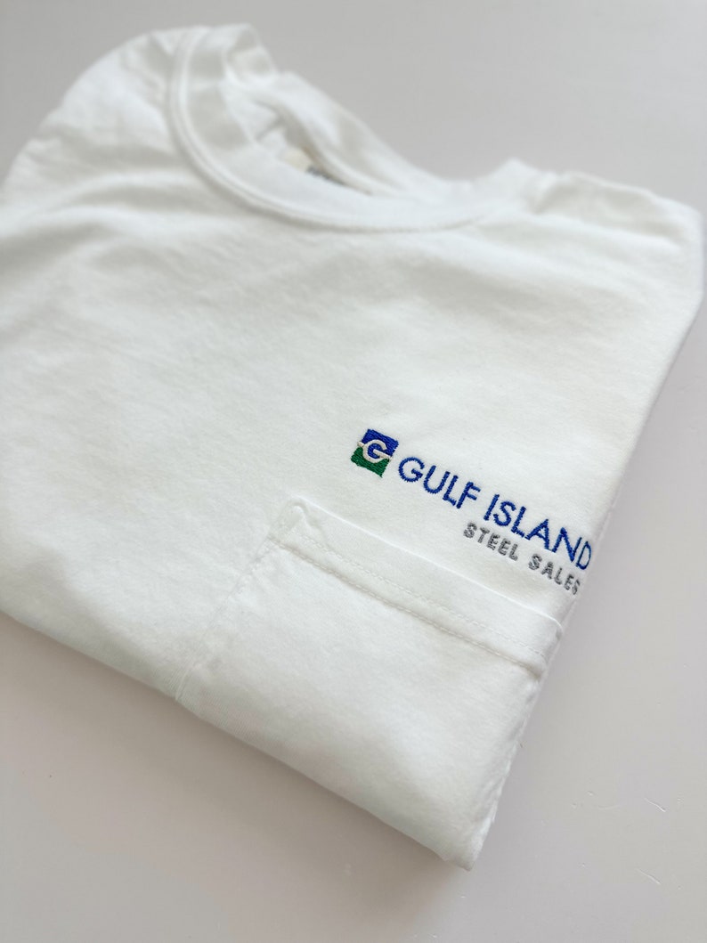Op de afbeelding: Wit T-shirt met een zak aan de linkerkant. De zak heeft een geborduurd logo met de tekst "GULF ISLAND STEEL SALES" met een groen, blauw en wit logo.