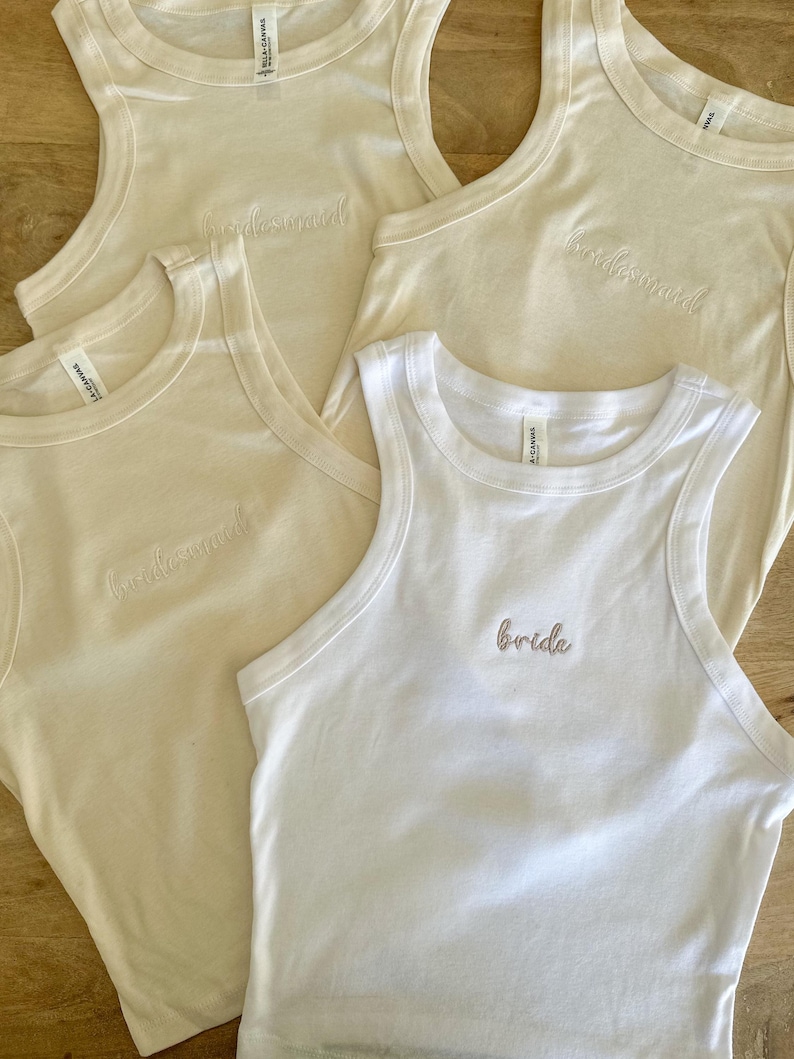 Pu&ograve; includere: Quattro canotte senza maniche color crema e bianche. Tre top color crema sono ricamati con la parola "bridesmaid" in corsivo. Una canotta bianca &egrave; ricamata con la parola "bride" in un carattere simile.