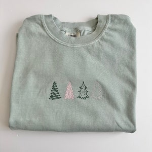 May include: A light green t-shirt with a row of four embroidered trees. The trees are in different colors, including green, pink, and black.