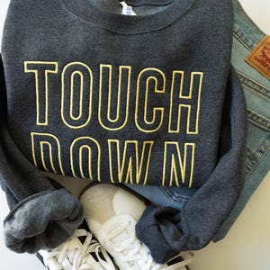 Puede incluir: Una sudadera gris oscuro con las palabras "TOUCH DOWN" en letras amarillas. La sudadera se combina con unos vaqueros azules y zapatillas blancas con detalles negros. Los artículos están colocados sobre una superficie blanca.