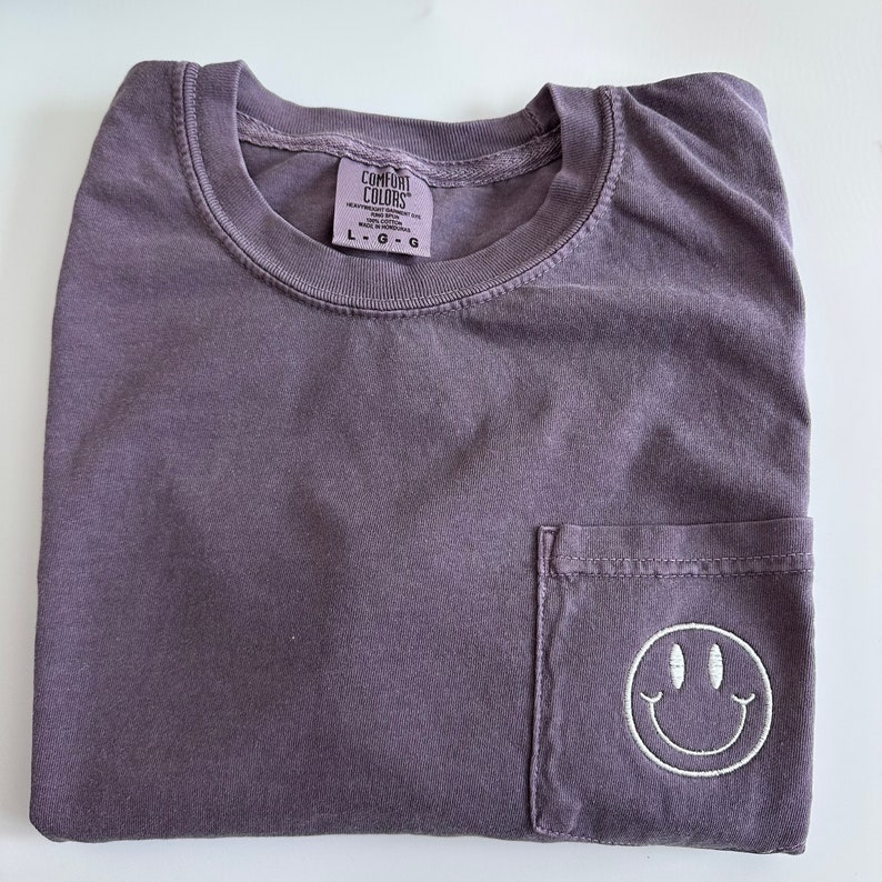 May include: A purple t-shirt with a pocket on the front. The pocket has a white embroidered smiley face. The t-shirt has a tag that says "Comfort Colors" and "L-G-G".