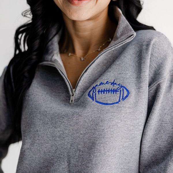 Könnte beinhalten: Ein graumeliertes Sweatshirt mit kurzem Reißverschluss und einer blauen Stickerei auf der linken Brust. Das Motiv zeigt die Umrisse eines Footballs mit dem Schriftzug "game day" in Schreibschrift direkt über der Football-Abbildung.