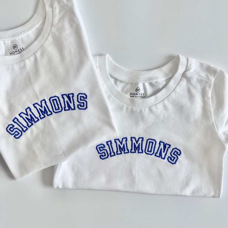 May include: Two white t-shirts with the name "SIMMONS" embroidered in blue. The shirts have a crew neck and the brand logo "HONEST" is visible on the collar. One shirt is larger than the other.