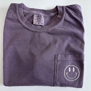 May include: A purple t-shirt with a pocket on the front. The pocket has a white embroidered smiley face. The shirt has a tag that says "Comfort Colors" and "L-G-G".