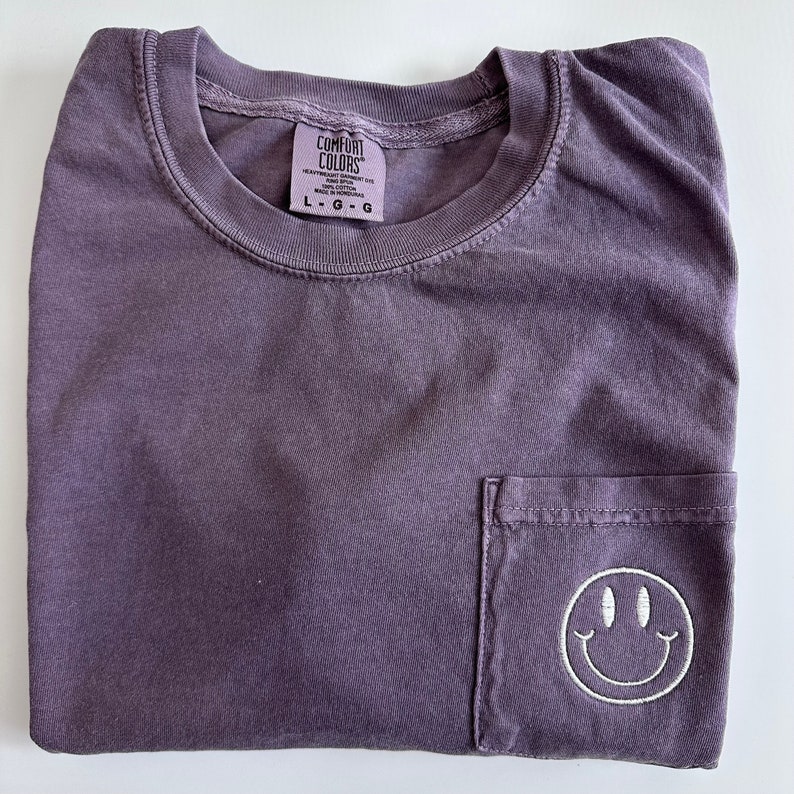 May include: A purple t-shirt with a pocket on the front. The pocket has a white embroidered smiley face. The shirt has a tag that says "Comfort Colors" and "L-G-G".
