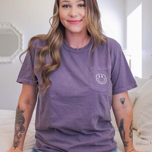 May include: A woman wearing a purple t-shirt with a white smiley face pocket and denim shorts. She has tattoos on her arms.