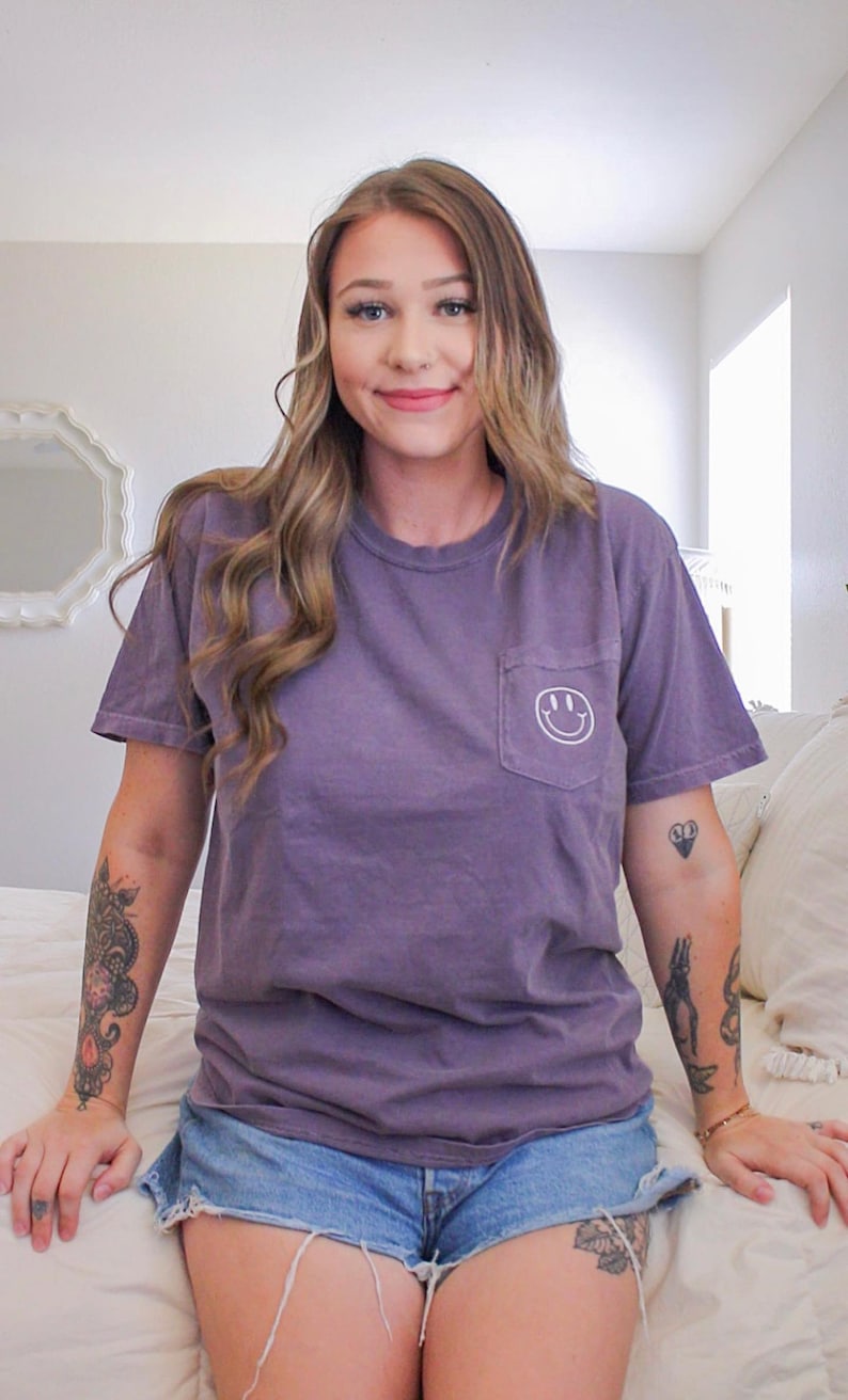May include: A woman wearing a purple t-shirt with a white smiley face pocket and denim shorts. She has tattoos on her arms.