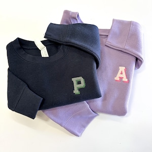 Puede incluir: Una sudadera azul marino oscuro con una letra P bordada en verde y gris, superpuesta a una sudadera lila claro con una letra A bordada en rosa y blanco. Ambas prendas tienen mangas largas y un cuello redondo.