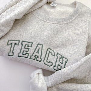 Op de afbeelding: Een grijs sweatshirt met het woord "TEACH" geborduurd in groen op de voorkant.