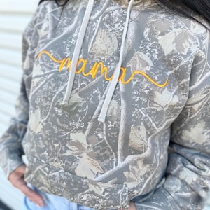 Könnte beinhalten: Ein Kapuzenpullover mit Camouflage-Muster und dem Wort "mama" in gelber Schrift auf der Brust gestickt. Der Kapuzenpullover hat eine Kapuze mit Kordelzug und lange Ärmel. Der Stoff hat ein graues, beiges und braunes Blättermuster.