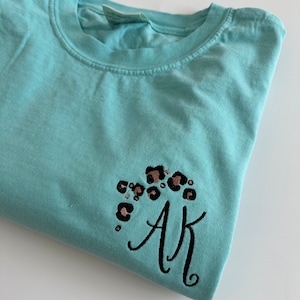 May include: A light turquoise crew neck t-shirt featuring an embroidered design on the lower right side. The embroidery consists of black and copper leopard print spots arranged in a cluster above the black cursive embroidered letters AK.