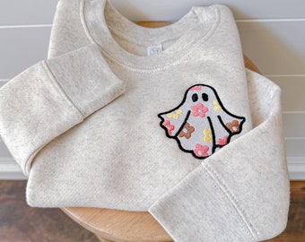 May include: A light beige heathered crewneck sweatshirt featuring an embroidered ghost graphic on the chest. The ghost is decorated with a pattern of small pink, yellow, and brown flowers and is outlined in thick black thread.