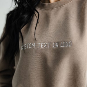 以下が含まれることがあります： 前面に白い刺繍で「CUSTOM TEXT OR LOGO」と刺繍された茶色のスウェットシャツです。