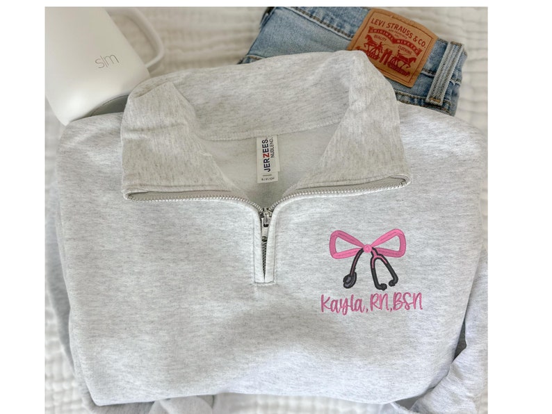 May include: A gray sweatshirt with a zipper and a pink bow with a stethoscope and the text "Kayla, RN, BSN" embroidered on the front.