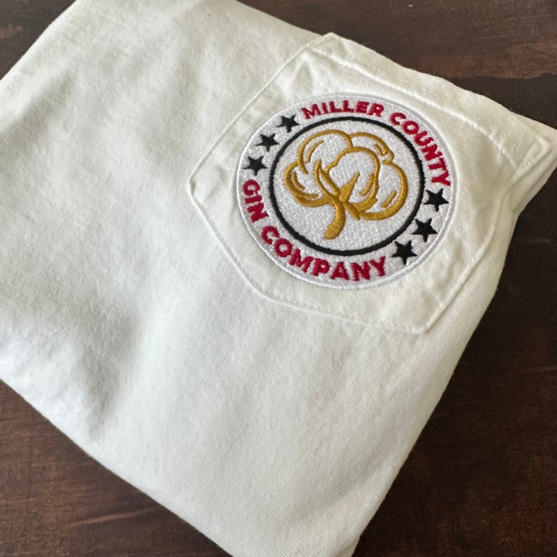 Op de afbeelding: Wit T-shirt met een geborduurde patch in rood, zwart en goud met de tekst "Miller County Gin Company" en een afbeelding van een katoenbol.