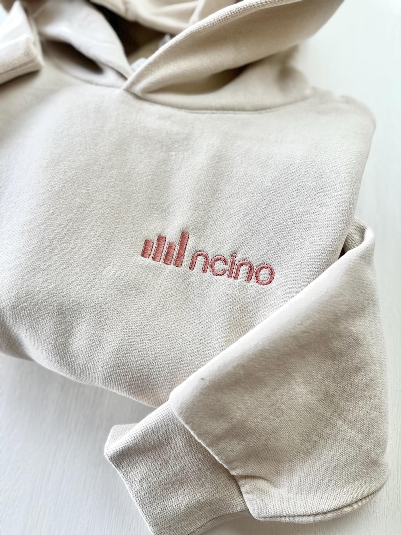 以下が含まれることがあります： フードと長袖が付いたベージュ色のパーカー。「Incino」の文字が赤茶色で前面に刺繍されています。パーカーは折りたたまれており、袖口が見えています。