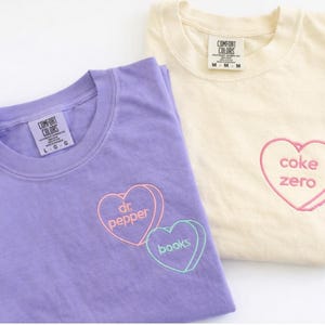 Może przedstawiać: Fioletowy t-shirt z wyhaftowanymi konturami serc zawierającymi tekst 'dr. pepper' i 'books' oraz kremowy t-shirt z wyhaftowanym konturem serca zawierającym tekst 'coke zero'. Oba t-shirty posiadają metki marki Comfort Colors.
