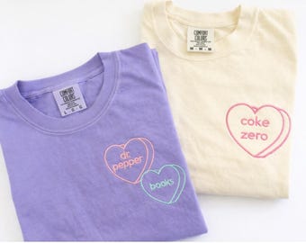 May include: A purple t-shirt with embroidered heart outlines containing the text 'dr. pepper' and 'books', and a cream t-shirt with an embroidered heart outline containing the text 'coke zero'. Both shirts feature Comfort Colors brand labels.