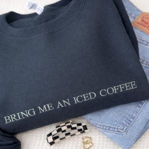 Könnte beinhalten: Ein schwarzes Sweatshirt mit Rundhalsausschnitt und dem Text BRING ME AN ICED COFFEE, der in hellgrauem Garn auf der unteren Vorderseite eingestickt ist. Das Sweatshirt liegt auf einer blauen Jeans und einer schwarz-weiß karierten Haarspange.