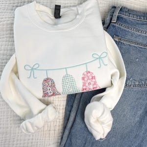 May include: A white crewneck sweatshirt featuring an embroidered design of three ghost shapes hanging from a light blue string with bows at each end. The ghosts are patterned with floral and gingham fabrics and display the letters B, O, and O.