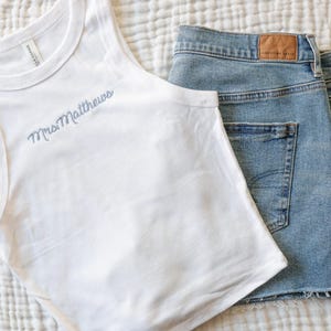 Pu&ograve; includere: Canotta bianca con la scritta ricamata "Mrs Matthews" in azzurro, abbinata a pantaloncini di jeans lavati chiari. I pantaloncini hanno un orlo grezzo e una toppa in pelle marrone.