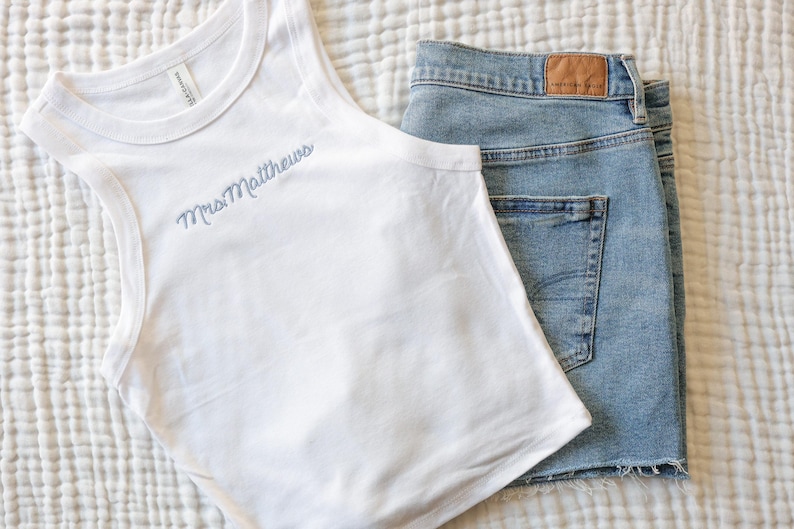Pu&ograve; includere: Canotta bianca con la scritta ricamata "Mrs Matthews" in azzurro, abbinata a pantaloncini di jeans lavati chiari. I pantaloncini hanno un orlo grezzo e una toppa in pelle marrone.