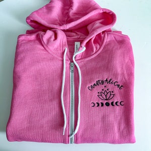 May include: A bright pink zip-up hoodie with a hood and white drawstrings. The front features black embroidery with the text Crafty Ali Cat above a lotus flower graphic and a series of moon phases. The garment has a silver-toned metal zipper.
