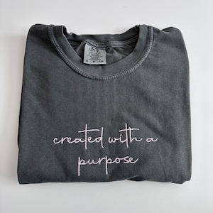 Puede incluir: Una camiseta gris oscura con el texto "created with a purpose" bordado en rosa en la parte delantera.