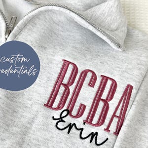 Puede incluir: Sudadera gris claro con cremallera de cuarto de cremallera. Cuenta con texto bordado burdeos y negro: "BCBA" y "Erin". Tambi&eacute;n es visible un c&iacute;rculo azul con el texto "custom credentials".