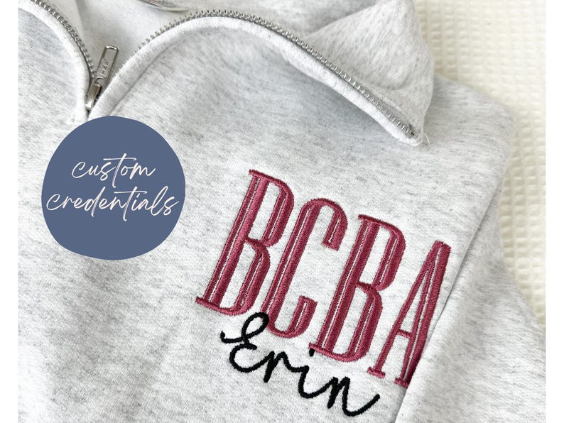 Puede incluir: Sudadera gris claro con cremallera de cuarto de cremallera. Cuenta con texto bordado burdeos y negro: "BCBA" y "Erin". Tambi&eacute;n es visible un c&iacute;rculo azul con el texto "custom credentials".