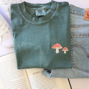 Pu&ograve; includere: Una maglietta verde acqua con un ricamo a forma di fungo rosso e bianco. La maglietta &egrave; appoggiata su un libro con un paio di scarpe da ginnastica bianche e un paio di jeans blu.