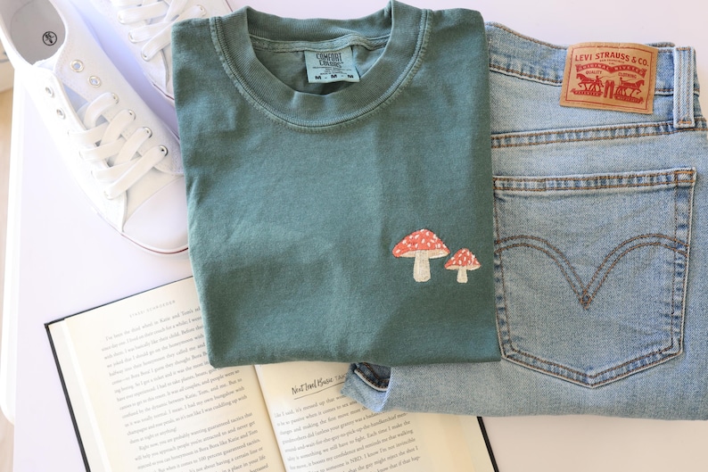 Pu&ograve; includere: Una maglietta verde acqua con un ricamo a forma di fungo rosso e bianco. La maglietta &egrave; appoggiata su un libro con un paio di scarpe da ginnastica bianche e un paio di jeans blu.