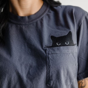 May include: Dark gray t-shirt with a pocket featuring an embroidered black cat design. The cat's eyes and whiskers are visible, adding a playful touch to the casual wear. The shirt is made of a soft material.