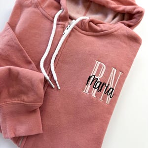 Puede incluir: Una sudadera con capucha de color rosa polvoriento con un cordón blanco y un texto bordado que dice "RN maria".