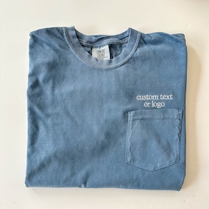 May include: A blue t-shirt with a pocket and the text "custom text or logo" embroidered on the pocket.