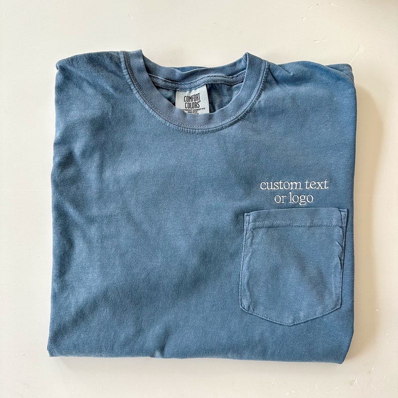 Op de afbeelding: Een blauw t-shirt met een zak en de tekst "custom text or logo" geborduurd op de zak.