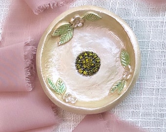 May include: A small, white ceramic dish with a raised floral design. The dish has a green and white floral pattern and a single green rhinestone in the center.