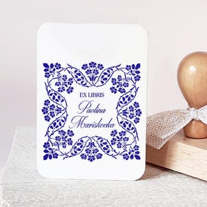 以下が含まれることがあります： 青い花の模様と「EX LIBRIS Paolina Marishooka」の文字が入った白い長方形のカード。明るい茶色のハンドルと白いリボンが付いた木製のスタンプがカードの隣にあります。スタンプは木のブロックの上にあります。