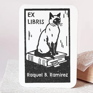 May include: A custom Ex Libris stamp featuring a black and white illustration of a cat sitting on a stack of books. The stamp includes the text "EX LIBRIS" and the name "Raquel B. Ramirez". The stamp has a wooden handle and base.
