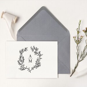 May include: A white card with a black floral wreath design and the initials "A M" is placed in front of a gray envelope. A spool of cream-colored ribbon and a sprig of flowers are also present.