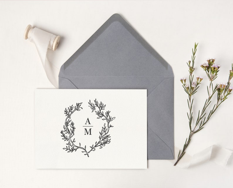 May include: A white card with a black floral wreath design and the initials "A M" is placed in front of a gray envelope. A spool of cream-colored ribbon and a sprig of flowers are also present.