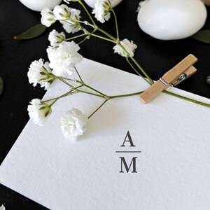 May include: A white card with the initials "A" over "M" is held by a wooden clothespin. White baby's breath flowers and white egg-shaped objects are scattered on a black surface. A minimalist design for invitations or announcements.