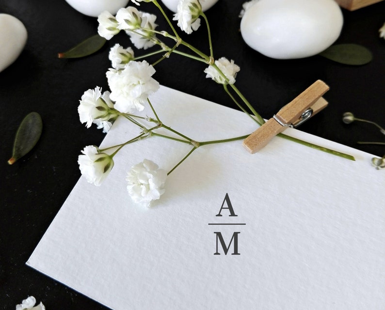 May include: A white card with the initials "A" over "M" is held by a wooden clothespin. White baby's breath flowers and white egg-shaped objects are scattered on a black surface. A minimalist design for invitations or announcements.