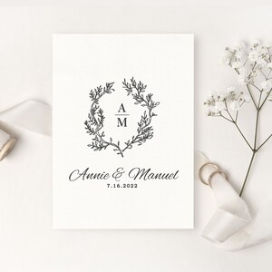 May include: A wedding invitation with a black floral wreath design, the initials "A" and "M", and the names "Annie & Manuel" with the date "7.16.2022". The invitation is on a white background, with a sprig of white flowers and two gold rings.