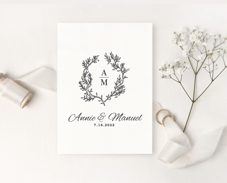 May include: A wedding invitation with a black floral wreath design, the initials "A" and "M", and the names "Annie & Manuel" with the date "7.16.2022". The invitation is on a white background, with a sprig of white flowers and two gold rings.